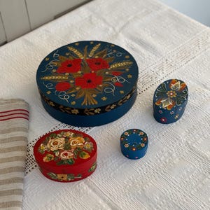 Op de afbeelding: Vier handgeschilderde houten dozen met bloemmotieven. De grootste doos is rond en blauw met rode en gele bloemen. De andere drie dozen zijn ovaal en hebben verschillende bloemmotieven in rood, geel en wit.