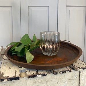 Puede incluir: Una bandeja redonda de metal marrón con asas sostiene un portavelas de cristal transparente y acanalado y hojas verdes. La bandeja descansa sobre una superficie blanca desgastada sobre un fondo blanco. El diámetro de la bandeja es de unos 30 cm.