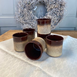 May include: Six brown and beige ceramic mugs with a speckled glaze. The mugs are arranged in a circle on a white tablecloth.
