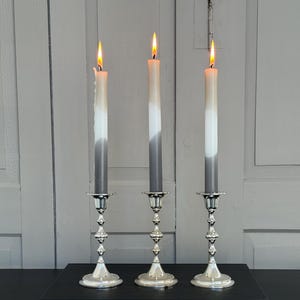 May include: Three silver candle holders with gray and white candles lit on a black surface.