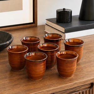 May include: A set of six small, cylindrical ceramic cups featuring a glossy, gradient finish that transitions from a deep, dark brown at the rim to a lighter, warm amber tone toward the base, with subtle horizontal ridges along the exterior surface.