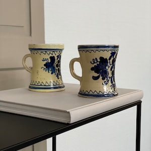 May include: Two vintage ceramic mugs with blue and white floral designs. The mugs have a cream-colored background and are sitting on a white book on a black metal table.