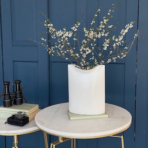 Könnte beinhalten: Weiße Keramikvase mit strukturierter Oberfläche, gefüllt mit einem Zweig weißer Blüten, auf einem Tisch mit weißer Marmorplatte und goldfarbenem Metallgestell.