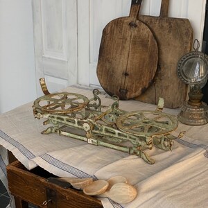 Pu&ograve; includere: Una bilancia vintage in metallo verde con due piattaforme rotonde e una leva. La bilancia &egrave; appoggiata su una tovaglia di lino bianco con strisce blu. Sullo sfondo ci sono due taglieri di legno e una lampada a olio di vetro.