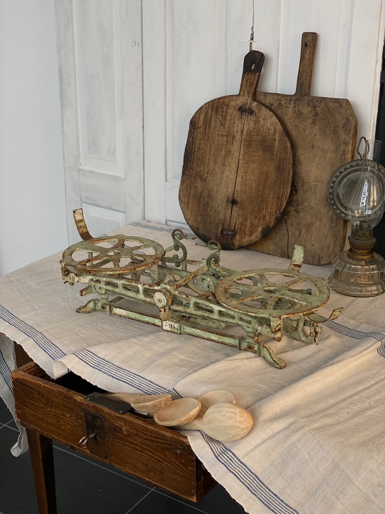 Pu&ograve; includere: Una bilancia vintage in metallo verde con due piattaforme rotonde e una leva. La bilancia &egrave; appoggiata su una tovaglia di lino bianco con strisce blu. Sullo sfondo ci sono due taglieri di legno e una lampada a olio di vetro.
