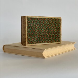 May include: A vintage book with a patterned cover in shades of green and orange. The book is sitting on top of a larger, plain book.