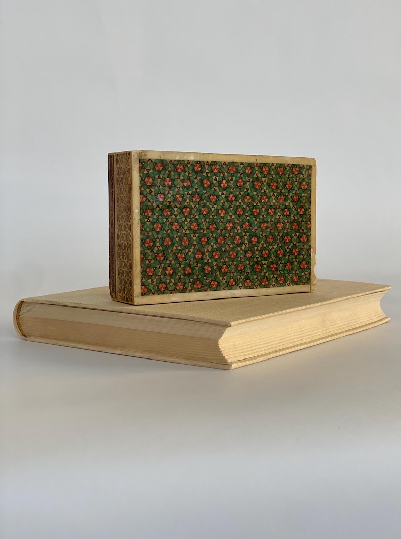 May include: A vintage book with a patterned cover in shades of green and orange. The book is sitting on top of a larger, plain book.