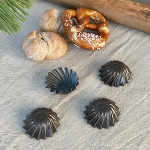 Könnte beinhalten: Vier schwarze Metall-Backformen mit geriffelten Rändern, angeordnet auf einer weißen Leinen-Tischdecke. Im Hintergrund ein Brezel und zwei Brötchen.