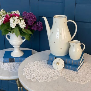 May include: A white ceramic teapot with a gold star pattern sits on a stack of blue books with a white crocheted doily on a white marble table. A white ceramic creamer sits next to the teapot. A white ceramic vase with a bouquet of pink and purple flowers sits on a similar table with a white crocheted doily.