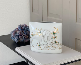 May include: White porcelain vase with a gold and white design of a woman and birds. The vase is sitting on a black shelf with books.