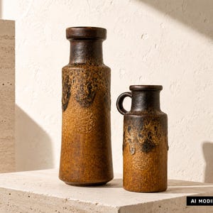 Könnte beinhalten: Zwei Keramikvasen mit einer texturierten, gesprenkelten Glasur in Braun und Dunkelschokolade. Die größere Vase ist zylindrisch mit einem ausgestellten Rand, während die kleinere Vase einen seitlichen Henkel und ein dunkles Tropfmuster besitzt.