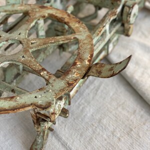 Pu&ograve; includere: Una bilancia vintage in metallo verde con una grande ruota circolare e una finitura arrugginita. La bilancia &egrave; appoggiata su una superficie di tessuto bianco.