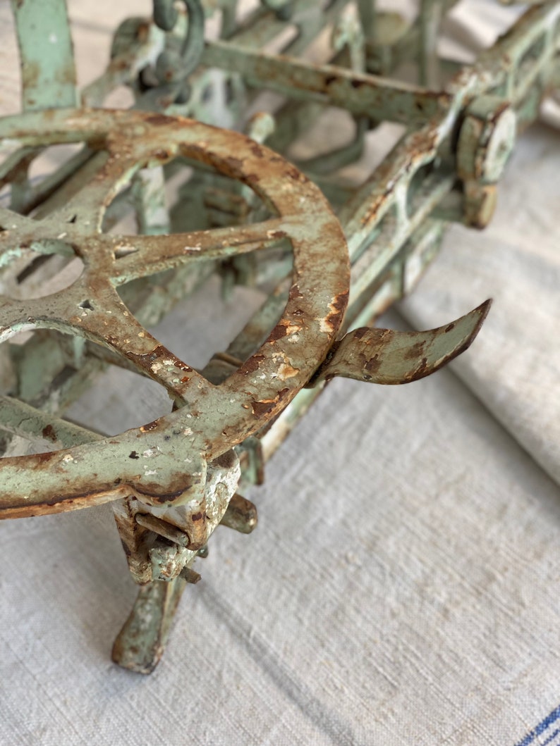 Pu&ograve; includere: Una bilancia vintage in metallo verde con una grande ruota circolare e una finitura arrugginita. La bilancia &egrave; appoggiata su una superficie di tessuto bianco.