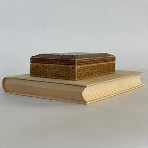 May include: A small, intricately patterned wooden box sits on top of a thick, off-white book. The box has a brown and gold geometric design.