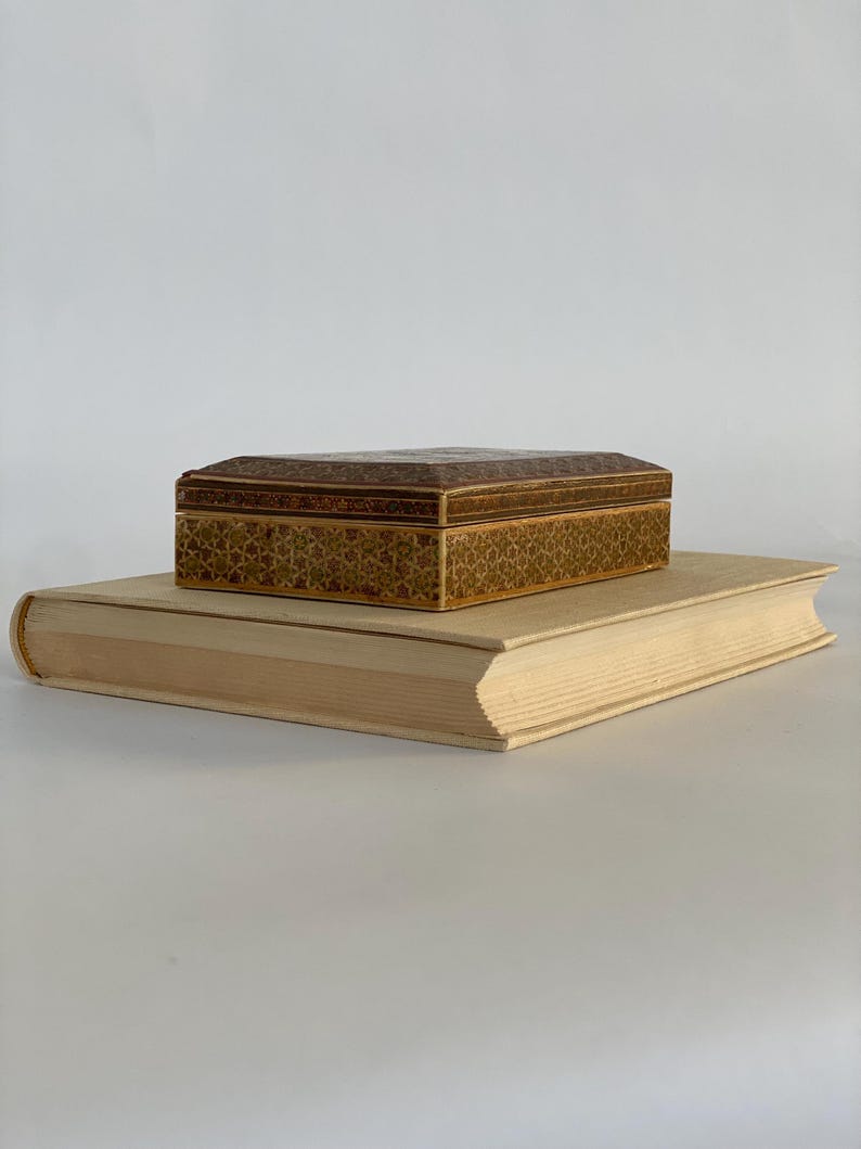 May include: A small, intricately patterned wooden box sits on top of a thick, off-white book. The box has a brown and gold geometric design.