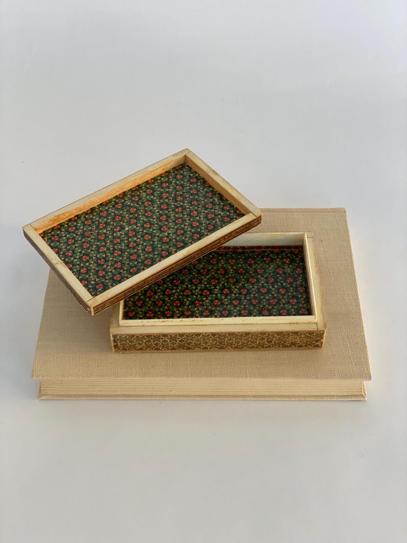 May include: A small wooden box with a floral pattern inside. The box is open and the lid is resting on top of a book.