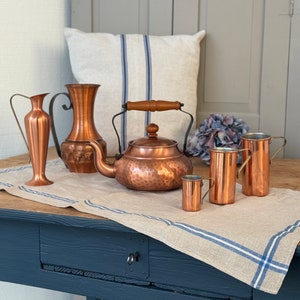 May include: A selection of copper kitchen items, including a kettle with a wooden handle, two vases, and small pitchers. They are arranged on a linen runner with blue stripes, placed on a dark blue table.