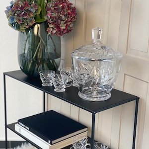 Könnte beinhalten: Ein schwarzes Metallregal mit zwei Ablagen. Die obere Ablage hat eine durchsichtige Glasbowle mit Deckel und vier kleine durchsichtige Gläser. Die untere Ablage hat ein schwarzes Buch mit weißem Text und vier weitere durchsichtige Gläser.