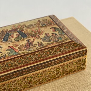 May include: A rectangular wooden box with intricate inlaid designs in shades of brown, green, and gold. The top of the box features a hand-painted scene of people and animals in a landscape setting.