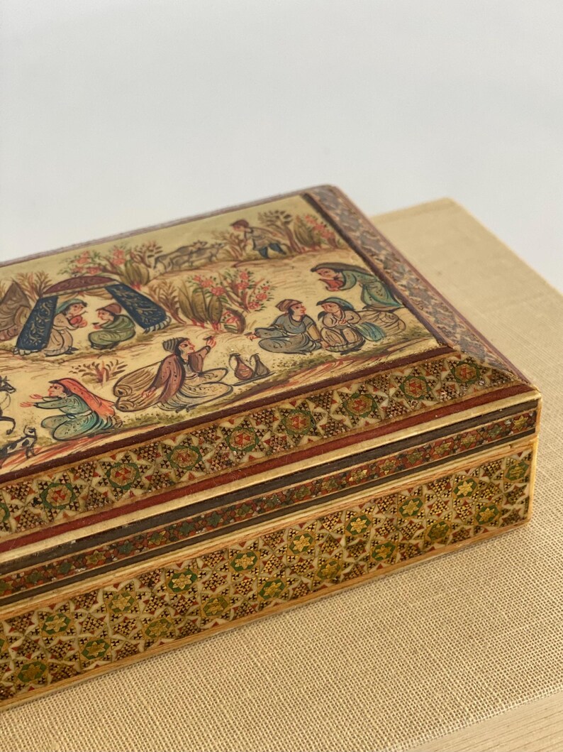 May include: A rectangular wooden box with intricate inlaid designs in shades of brown, green, and gold. The top of the box features a hand-painted scene of people and animals in a landscape setting.