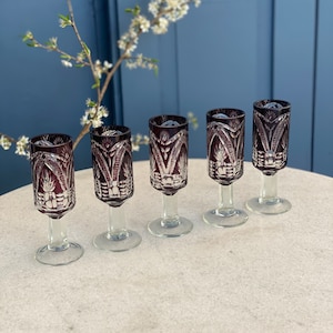 Puede incluir: Conjunto de cinco vasos de chupito de vidrio transparente con un tono morado intenso y diseños intrincados de vidrio cortado. Los vasos tienen un tallo corto y una base redonda.