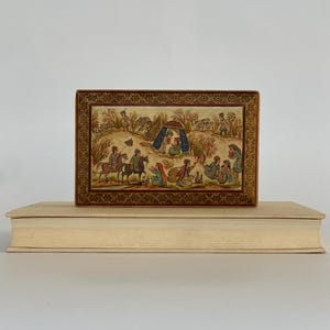 May include: A small wooden box with a hinged lid, decorated with intricate gold and brown patterns. The lid features a hand-painted scene of people on horseback and a tent in a landscape.
