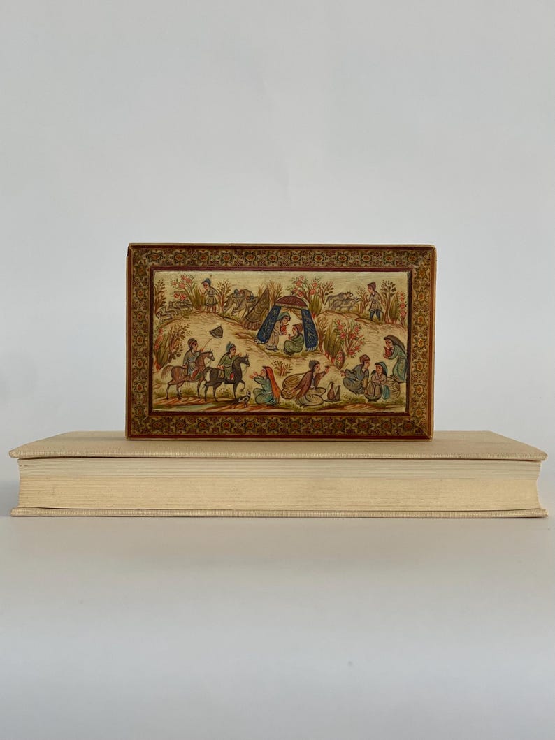 May include: A small wooden box with a hinged lid, decorated with intricate gold and brown patterns. The lid features a hand-painted scene of people on horseback and a tent in a landscape.