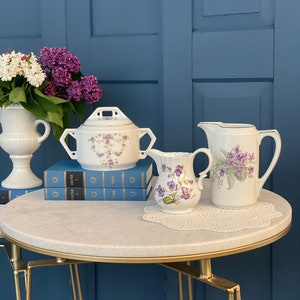 May include: A white porcelain sugar bowl with a lid and floral design, a white porcelain creamer with a floral design, and a white porcelain pitcher with a floral design, all sitting on a round marble table with a gold metal base.
