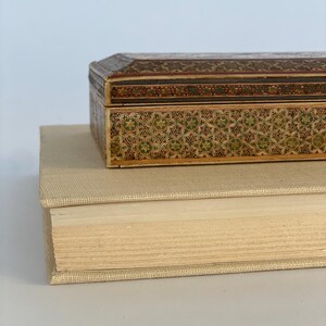 May include: A small, intricately carved wooden box with a geometric pattern in shades of brown, green, and gold. The box is sitting on top of a thick, cream-colored book.
