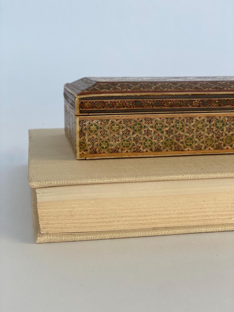 May include: A small, intricately carved wooden box with a geometric pattern in shades of brown, green, and gold. The box is sitting on top of a thick, cream-colored book.