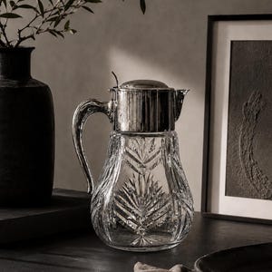 Könnte beinhalten: Ein Krug aus klarem Glas mit einem kunstvollen Schliffmuster in Form von palmblattartigen Motiven. Der Krug verfügt über einen polierten, silberfarbenen Metalldeckel mit einem kleinen Hebel sowie einen passenden silberfarbenen Henkel.