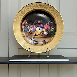 Könnte beinhalten: Ein dekorativer Teller mit einem Blumenkorb-Design. Der Teller hat einen goldenen Rand mit aufwendigen Details und ein zentrales Bild eines Korbes voller bunter Blumen. Der Teller wird auf einem schwarzen Ständer präsentiert.