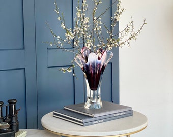 May include: A white and purple glass vase with a ruffled top, filled with white blossoms, sits on a round marble table with gold legs. The table is next to a blue wall.