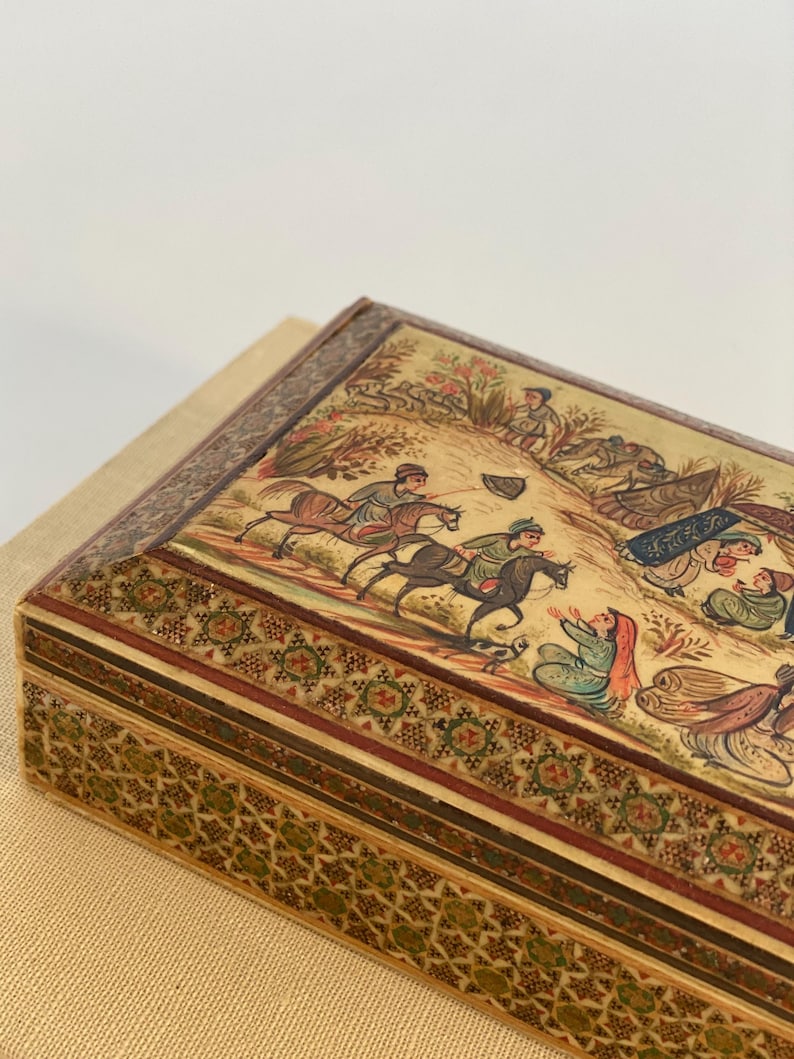 May include: A rectangular wooden box with intricate inlaid designs in shades of brown, green, and gold. The top of the box features a hand-painted scene depicting people on horseback and figures in a landscape.
