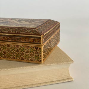 May include: A small, intricately decorated wooden box with a floral pattern and a geometric design on the sides. The box is sitting on top of a thick, cream-colored book.