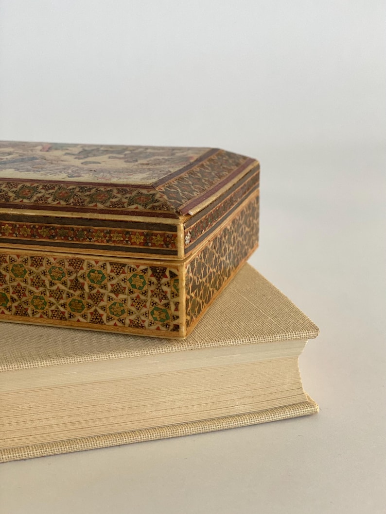 May include: A small, intricately decorated wooden box with a floral pattern and a geometric design on the sides. The box is sitting on top of a thick, cream-colored book.