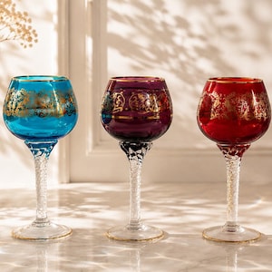 Op de afbeelding: Drie glazen op voet in turquoise, dieppaars en rood, elk voorzien van een gouden filigraanband rond de kelk en een heldere, gedraaide glazen steel. De randen en ronde voeten zijn afgewerkt met een dunne gouden bies, wat een set vormt.