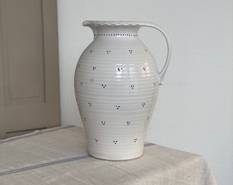 May include: White ceramic pitcher with a blue polka dot pattern and a scalloped rim. The pitcher has a handle and is sitting on a white tablecloth with blue stripes.