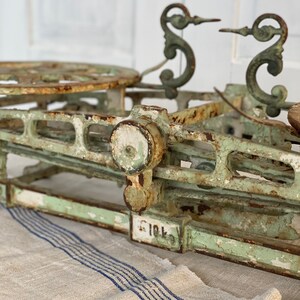 Pu&ograve; includere: Bilancia antica in ghisa verde con accenti in metallo decorativi e un quadrante bianco. La bilancia &egrave; appoggiata su un tessuto a strisce bianche e blu.