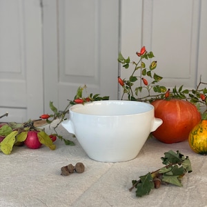 May include: White ceramic bowl with two handles, sitting on a white linen tablecloth. The bowl is surrounded by fall foliage, including red berries, acorns, and a small orange pumpkin.