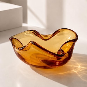 Könnte beinhalten: Bernsteinfarbene Glasschale mit wellenförmiger, organischer Form. Die Schale hat ein breites, offenes Design mit glatter, glänzender Oberfläche. Das Glas ist durchscheinend, wodurch Licht hindurchscheinen und Schatten erzeugen kann. Die Schale wird wahrscheinlich zu dekorativen Zwecken verwendet.