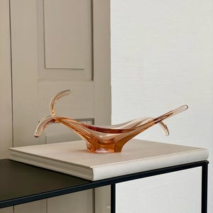 May include: A peach-colored glass bowl with a unique, flowing design. The bowl has two upward-curving extensions on either side. It sits on a white book, placed on a black metal table. The background includes a light-colored door and a textured white wall.
