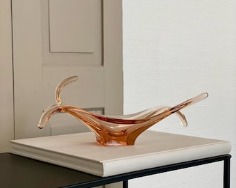 Könnte beinhalten: Eine pfirsichfarbene Glasschale mit einem einzigartigen, fließenden Design. Die Schale hat zwei nach oben gebogene Verlängerungen auf beiden Seiten. Sie steht auf einem weißen Buch, das auf einem schwarzen Metalltisch liegt. Der Hintergrund umfasst eine hellfarbige Tür und eine strukturierte weiße Wand.