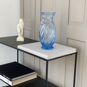 May include: A blue glass vase with a swirl design sits on a white book on a black metal shelf. A white sculpture of two faces is on the shelf below.