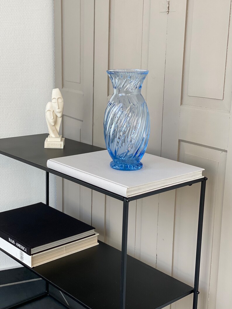 May include: A blue glass vase with a swirl design sits on a white book on a black metal shelf. A white sculpture of two faces is on the shelf below.