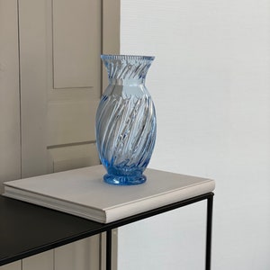 May include: A light blue glass vase with a swirl design sits on a white book on a black metal table.