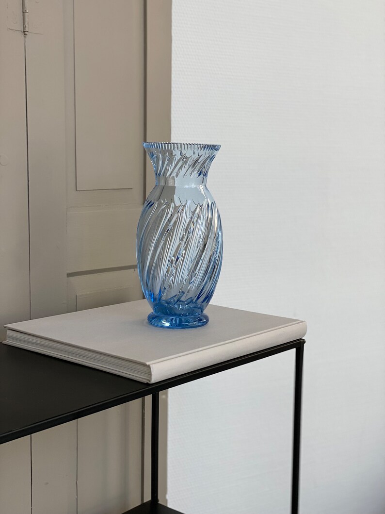 May include: A light blue glass vase with a swirl design sits on a white book on a black metal table.