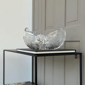 Puede incluir: Un cuenco de cristal transparente con un diseño grabado de uvas y hojas. El cuenco está sobre un libro negro, que a su vez está sobre una estantería de metal negro. La estantería está contra una pared blanca.