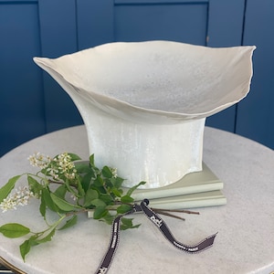 May include: A white ceramic vase with a unique, organic shape. The vase is sitting on a marble table with a green plant and a brown ribbon with the text "Hermes - Paris" on it.