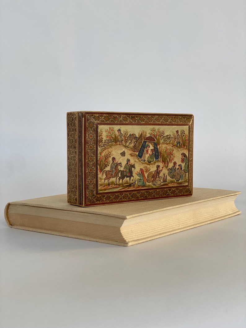 May include: A small wooden box with a hinged lid. The box is decorated with intricate floral patterns and a painted scene of people on horseback and a group of people gathered under a tent. The box is a traditional Persian style.